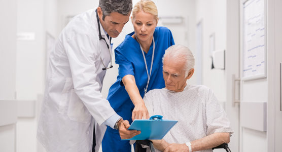 Doctor and nurse with senior patient in wheelchair at hospital corridor talking. Mature doctor showing and discussing medical report with nurse and senior disabled at medical clinic.